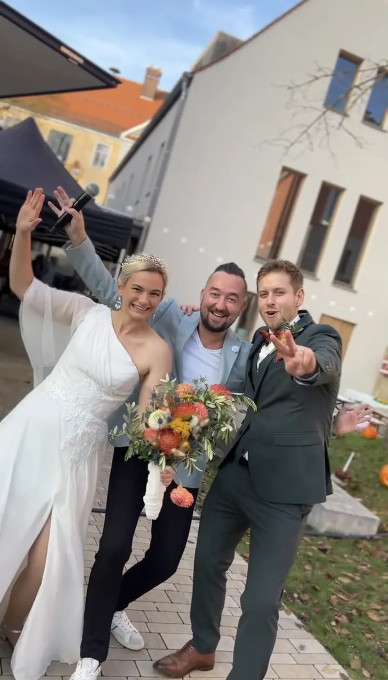 Braut mit orangem Brautstrauß, Bräutigam und Moderator vor weißem Gebäude Braut mit orangem Brautstrauß, Bräutigam und Moderator vor weißem Gebäude