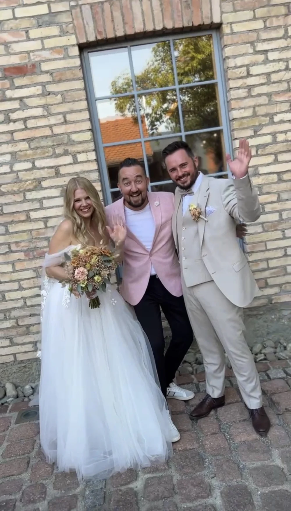 Braut, Bräutigam und Moderator Andrey Singer im rosa Sakko vor einer Backsteinwand Braut, Bräutigam und Moderator Andrey Singer im rosa Sakko vor einer Backsteinwand