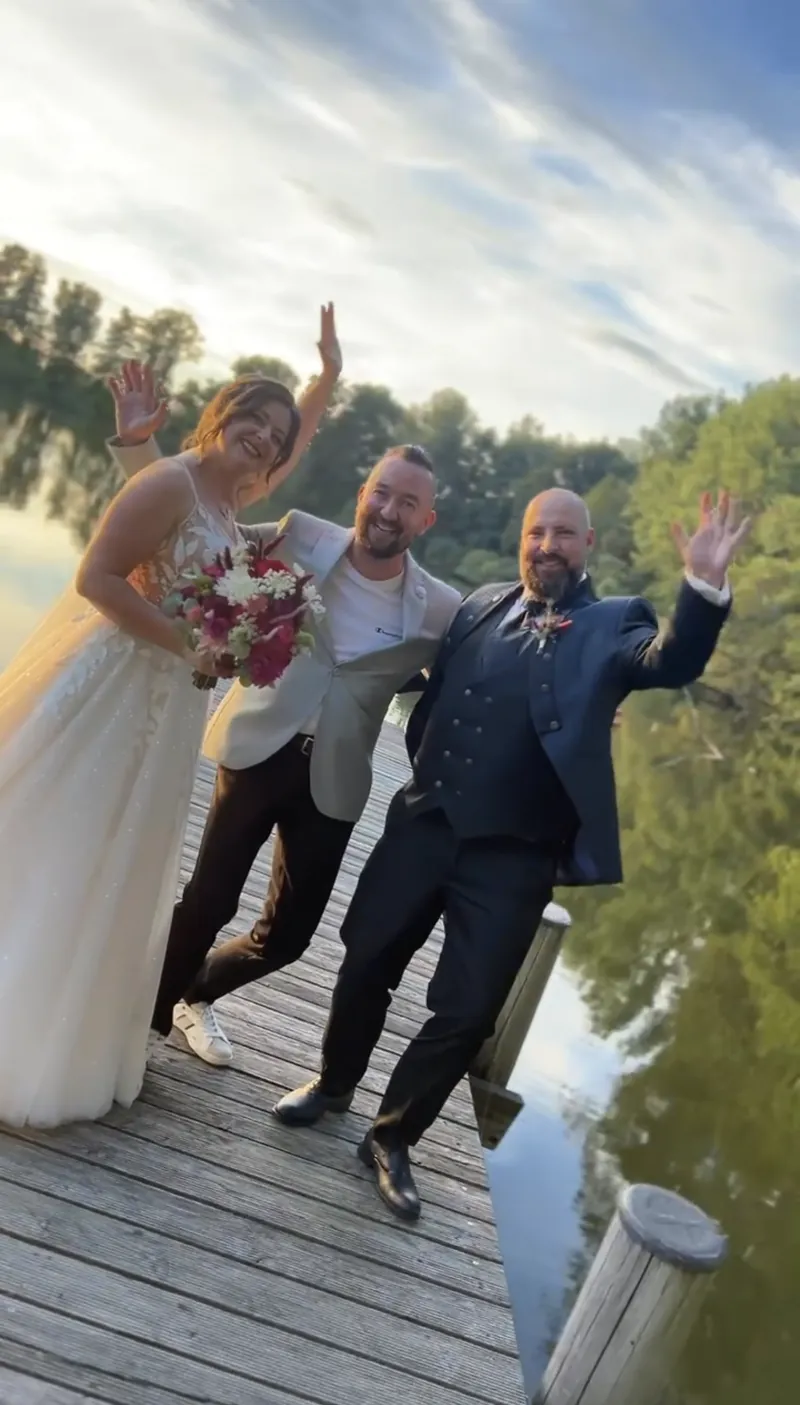 Brautpaar und Hochzeitsmoderator winken auf einem Holzsteg am See Brautpaar und Hochzeitsmoderator winken auf einem Holzsteg am See